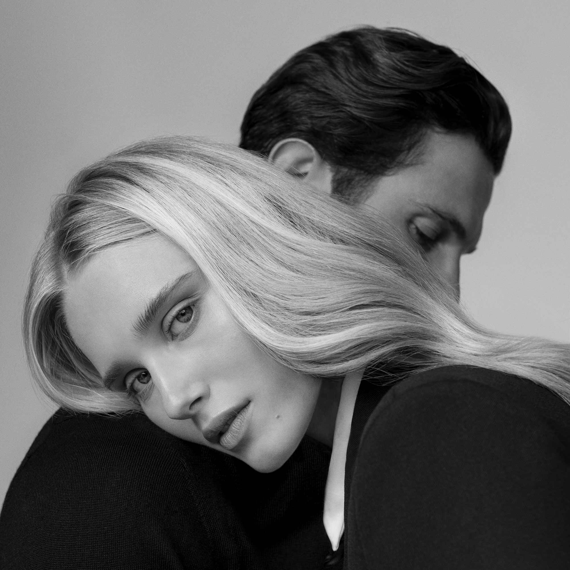black and white photo of woman with beautiful blonde hair leaning on the shoulder of a brunette man and looking at the camera